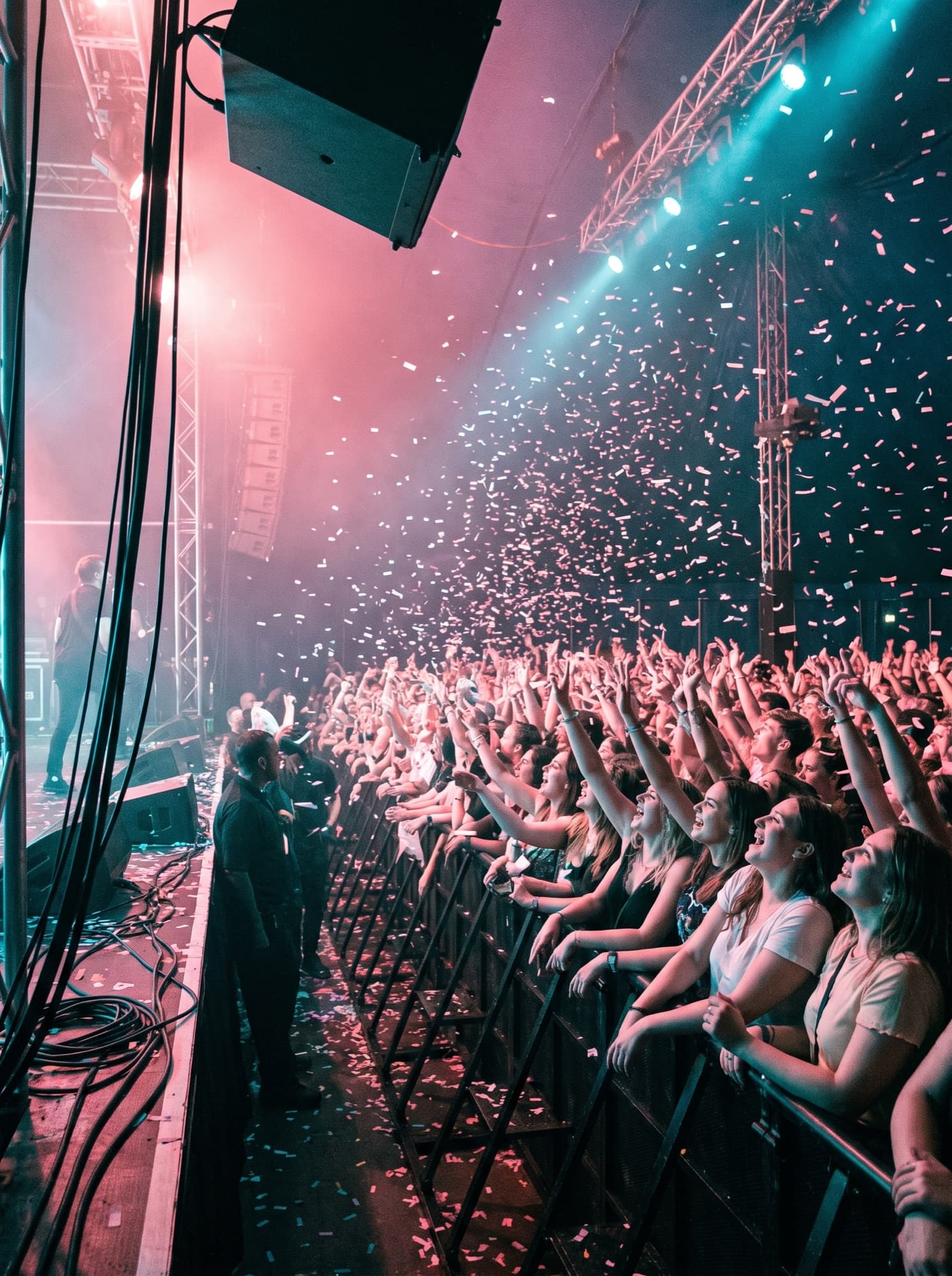 The front rows of a live show, arms raised under pink and cyan light